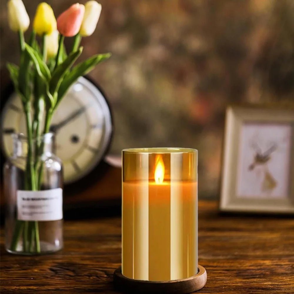 Candle in a glass holder on a wooden surface with flowers and a clock in the background